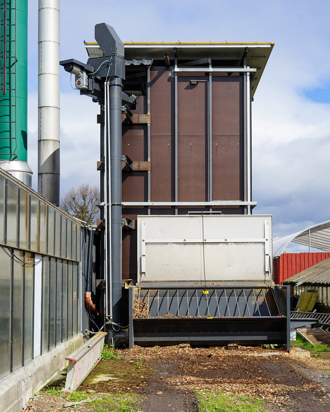 senkrechtfoerderer-eindrueckschnecke-bunkerbefuellung Brauner Brennstoffbunker aus Siebdruckplatten mit Fördertechnik vor einem Gewächshaus