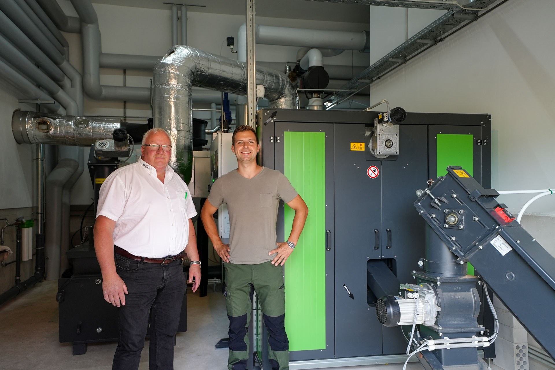 referenz-blandau-ak1400 Zwei Personen vor einer grünen Hackschnitzelheizung von Heizomat