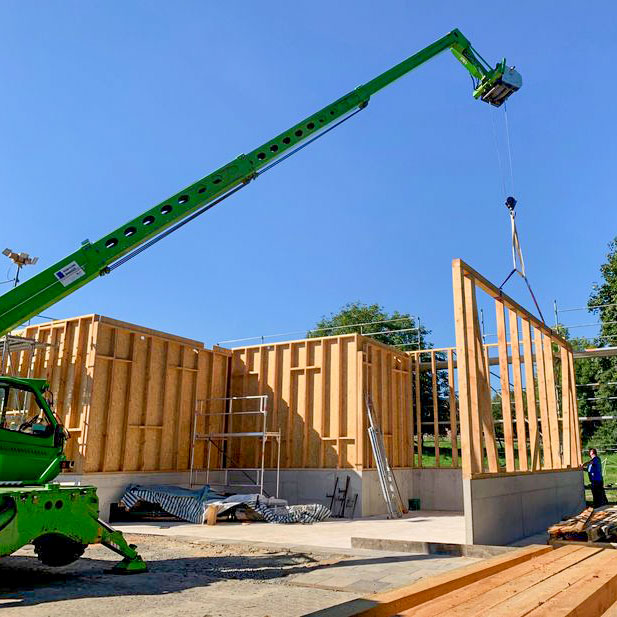 richten-holzhalle-heizzentrale Ein grüner Merlo setzt mit seinem Arm ein Holzbauelement auf einen Betonsockel