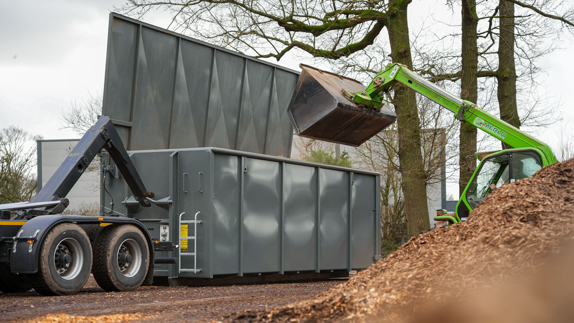 hakenliftanhänger-für-container Merlo beim Befüllen eines grauen Containers mit Hackschnitzeln